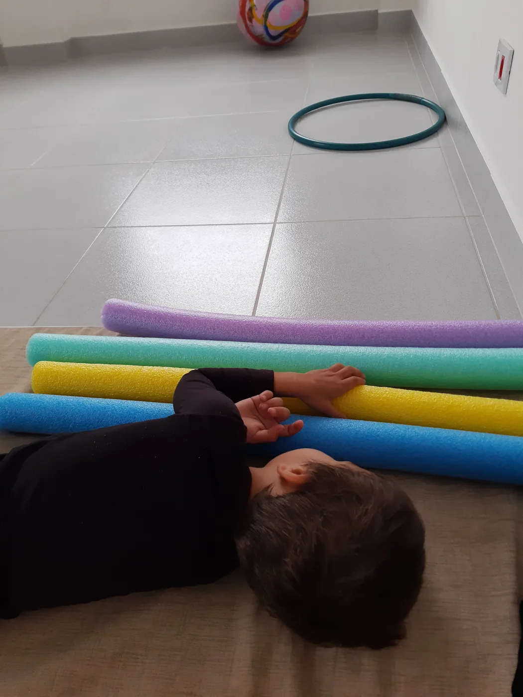 Criança deitada de bruços no tapete, com os braços apoiados sobre espaguetes de espuma coloridos; ao fundo, um bambolê e uma bola no chão do salão