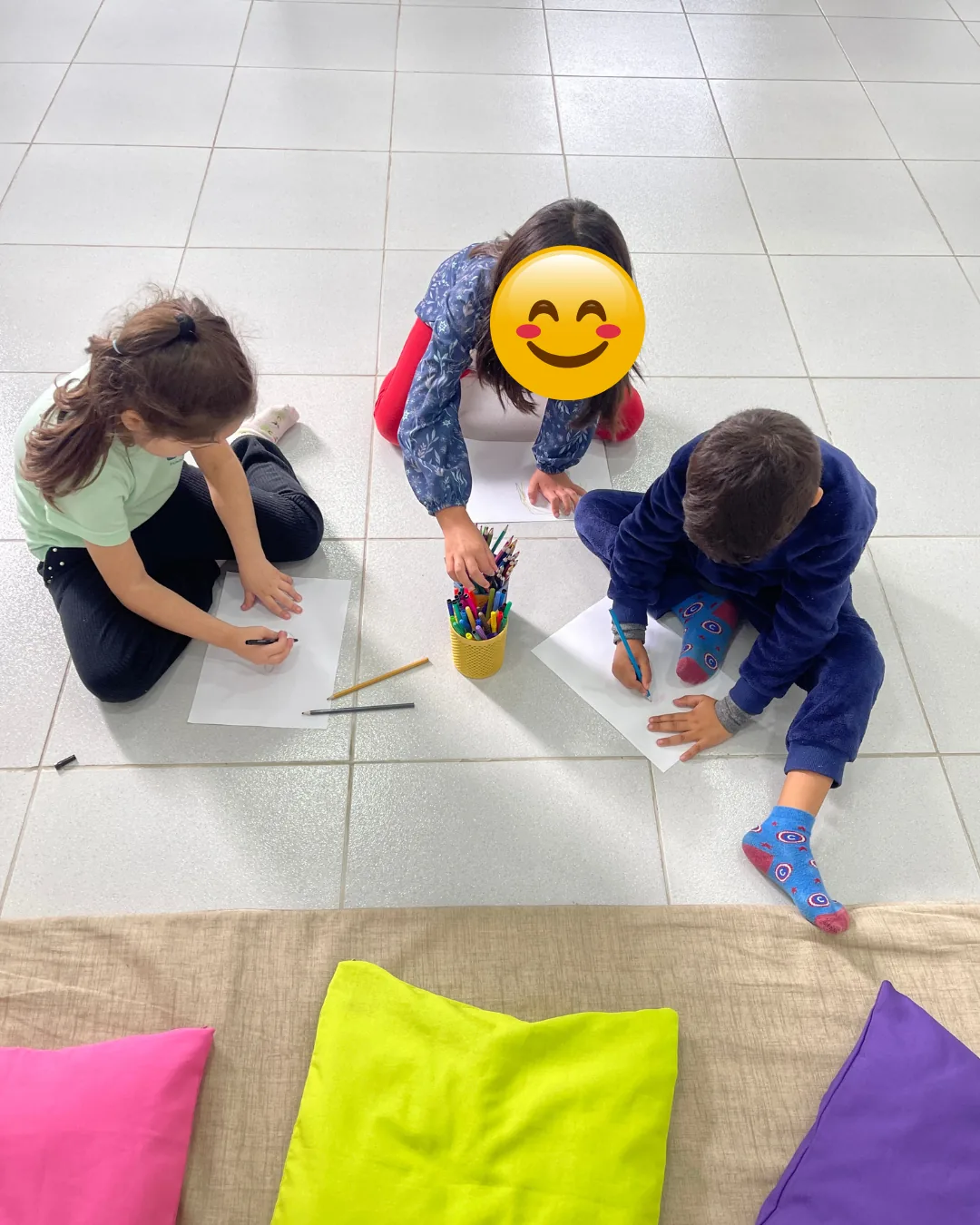 Três crianças sentadas no chão do salão desenhando em folhas de papel, com um pote de canetinhas coloridas entre elas e almofadas ao lado