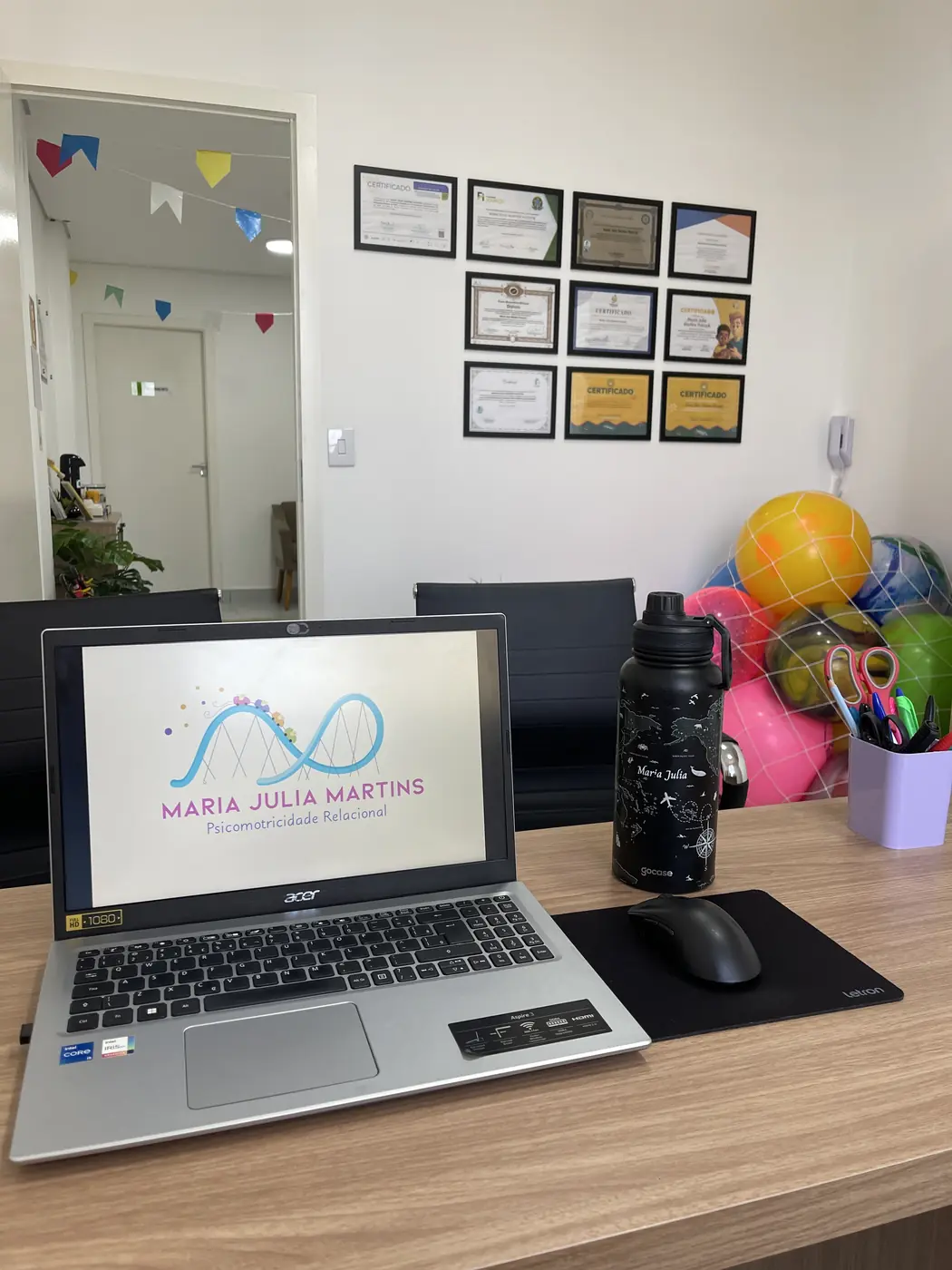 Mesa do escritório com notebook exibindo a logo Maria Julia Martins e certificados de formação na parede